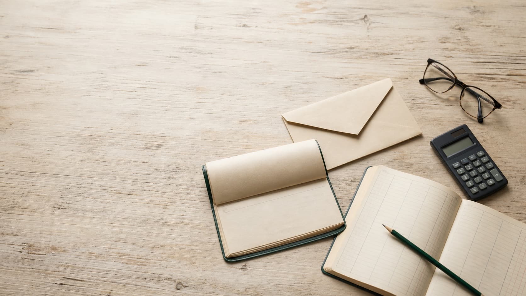 A blank ledger, envelope, calculator, glasses, and pencil arranged on a wooden desk