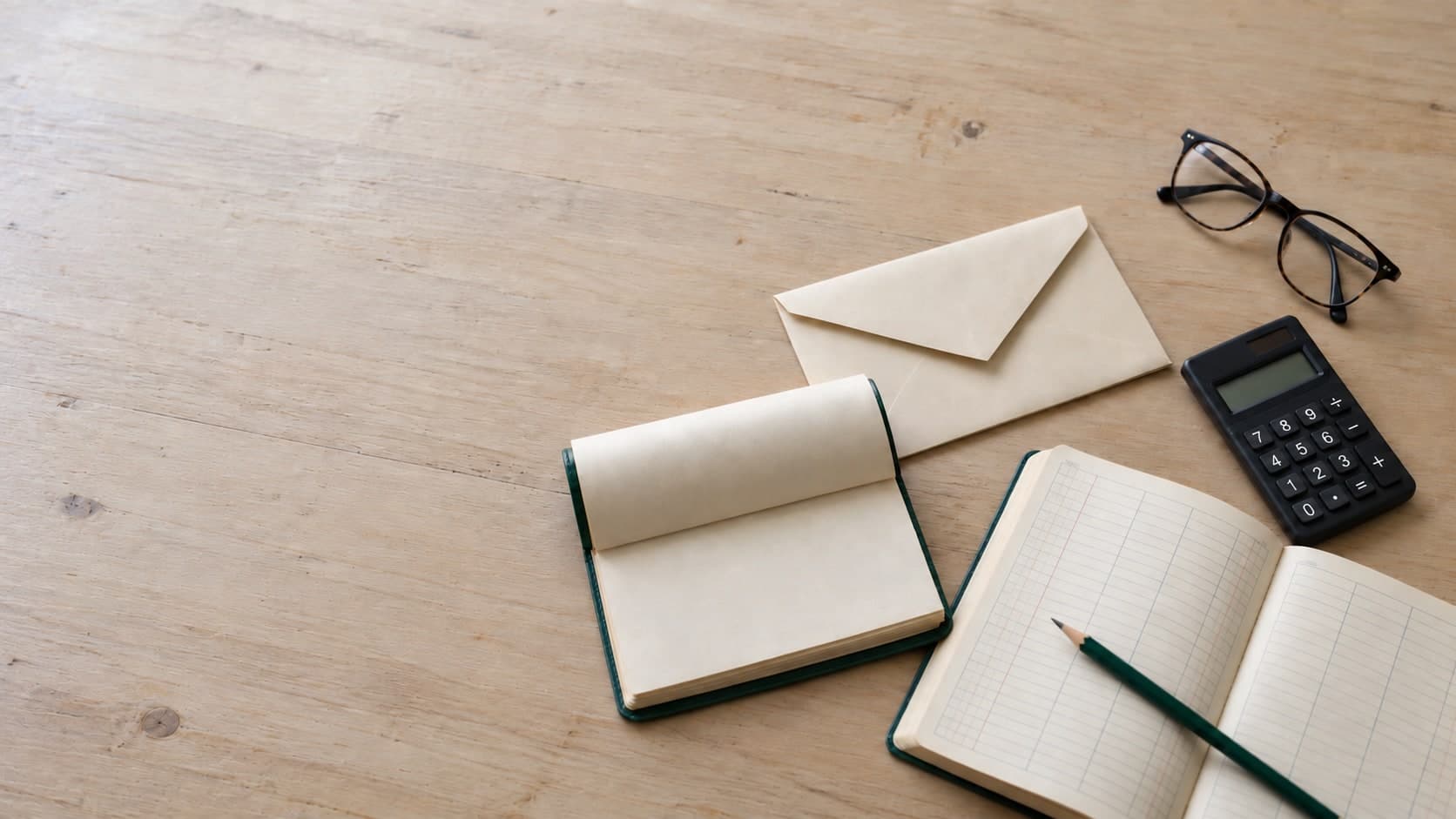 Blank notebooks, an envelope, calculator, glasses, and pencil on a wooden desk